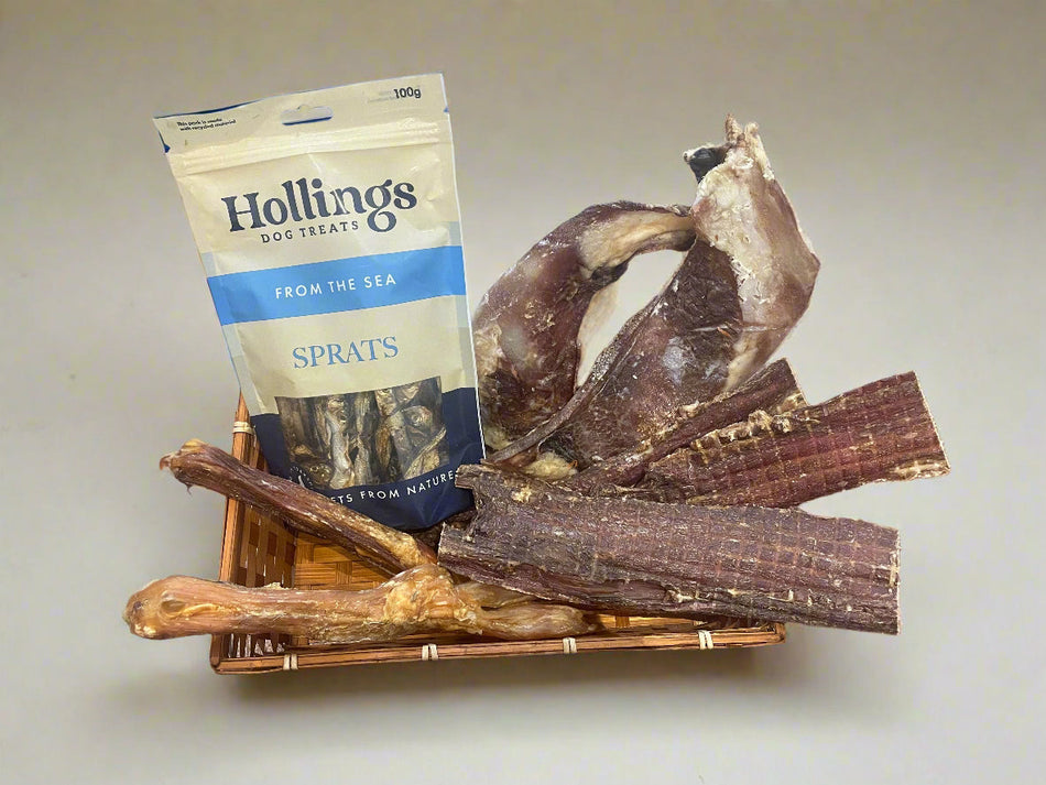 The Ultimate Natural Treat Bundle For Dogs featuring Hollings Sprats, biltong, moon bones, and beef tendons in a wicker basket for healthy dog snacking.