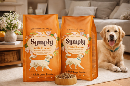 Two bags of Symply Large Breed Chicken dog food for adult large breeds with a dog sitting next to them in a living room.